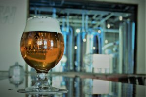 Belgian style footed glass with Adventurepants Double IPA. Blurry background with colorful brewhouse.
